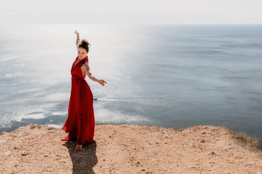 Side view a Young beautiful sensual woman in a red long dress posing on a rock high above the sea during sunrise. Girl on the nature on blue sky background. Fashion photo.