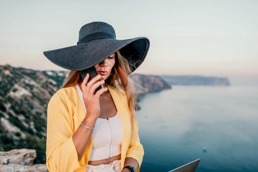 Successful business woman in yellow hat working on laptop by the sea. Pretty lady typing on computer at summer day outdoors. Freelance, travel and holidays concept.