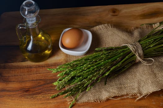 bunch of wild asparagus on a raffia cloth with a jar of olive oil and eggs black background