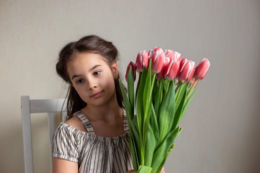 Portrait of a beautiful romantic little girl with a bouquet of pink tulips, sits on a chair, against a gray wall, looks away. copy space