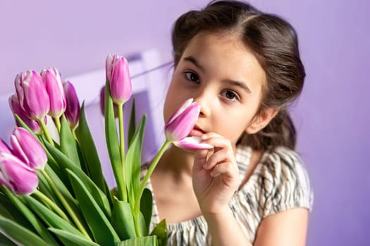 Portrait of a beautiful little girl with a bouquet of tulips, sniffs the aroma of a bud, looks at the camera, purple background. Close up