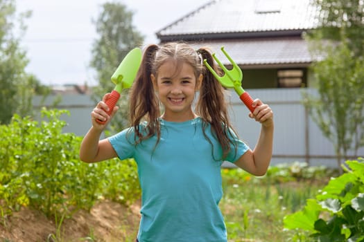 Cute little girl in green t-shirt holding small gardening tools , standing in the garden on a sunny day. Copy space