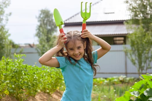 Funny little girl in green t-shirt holding small gardening tools , standing in the garden on a sunny day. Copy space