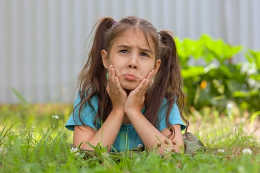 Sad brunette girl with two tails lies on the green grass, on her stomach, in the summer garden, holding thumb up. Copy space