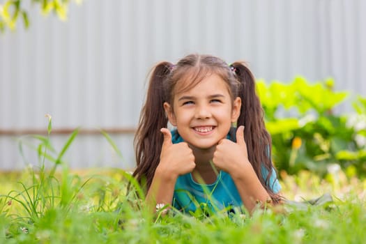 Cute smiling brunette girl with two tails lies on the green grass, on her stomach, in the summer garden, holding two thumbs up