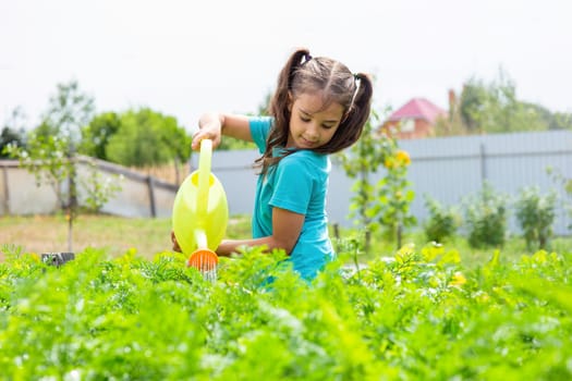 Smiling little girl in a green T-shirt, watering a green bed of yellow watering can, in the garden, on a sunny day. Copy space. Close up
