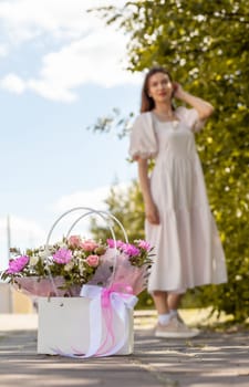 A beautiful bouquet of flowers in a box in the hands of a beautiful girl who walks along the street on a sunny day. Girl in a dress, glasses and sneakers. Focus on the background of flowers.