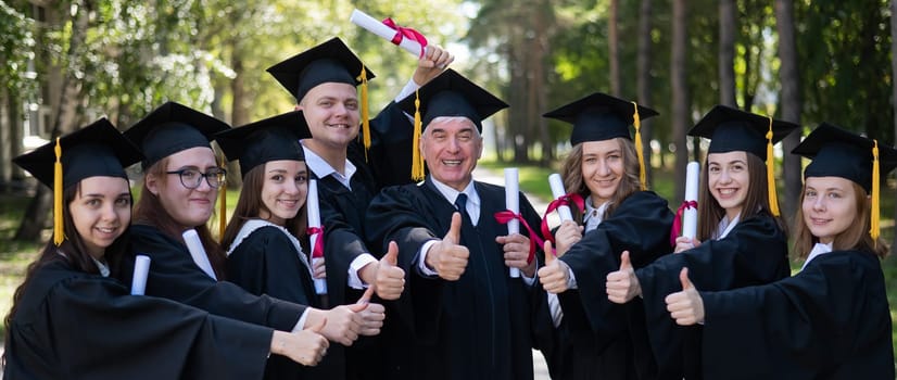 A group of graduates in robes give a thumbs up outdoors. Elderly student