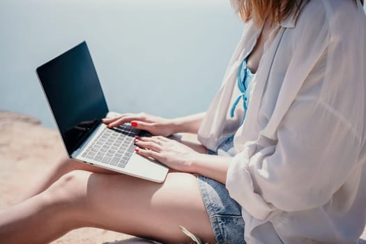 Successful business woman in yellow hat working on laptop by the sea. Pretty lady typing on computer at summer day outdoors. Freelance, travel and holidays concept.