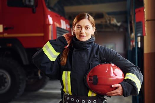 Holds red hat in hands. Female firefighter in protective uniform standing near truck.