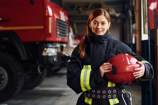 Holds red hat in hands. Female firefighter in protective uniform standing near truck.