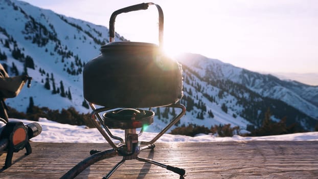 The burner is burning with a blue fire with a kettle. The burner and the cylinder are on the bench. In the background there are green fir trees, mountains in white snow and a beautiful sunset