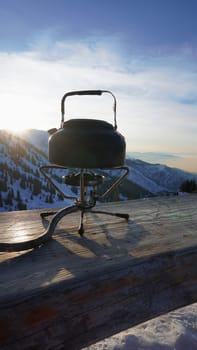 The burner is burning with a blue fire with a kettle. The burner and the cylinder are on the bench. In the background there are green fir trees, mountains in white snow and a beautiful sunset