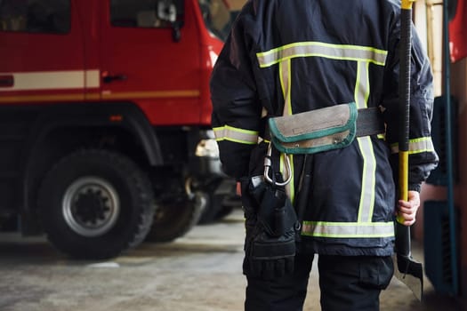 Holds axe in hands. Male firefighter in protective uniform standing near truck.