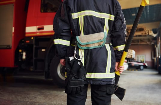 Holds axe in hands. Male firefighter in protective uniform standing near truck.