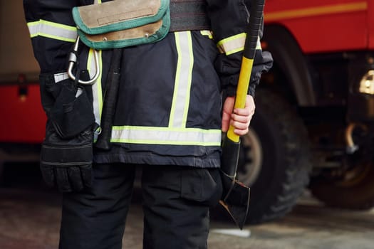Holds axe in hands. Male firefighter in protective uniform standing near truck.