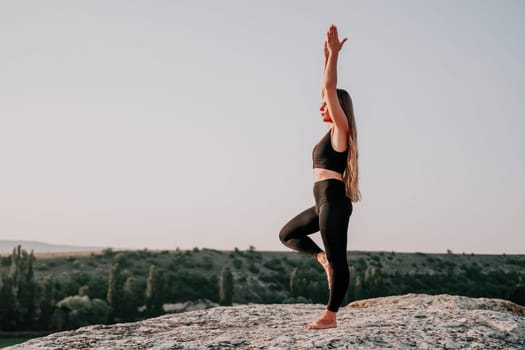 Well looking middle aged woman with long hair, fitness instructor in leggings and tops doing stretching and pilates on the rock near forest. Female fitness yoga routine concept. Healthy lifestyle.
