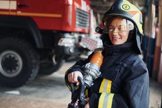 Hose in hands. Female firefighter in protective uniform standing near truck.