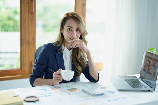 Portrait of an Asian business woman drinking coffee while working with a computer and financial statements documents on her desk.