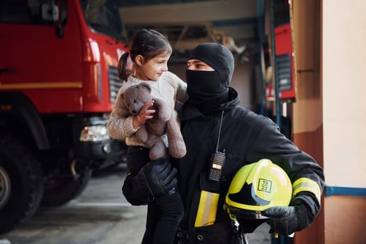 Holds teddy bear. Happy little girl is with male firefighter in protective uniform.