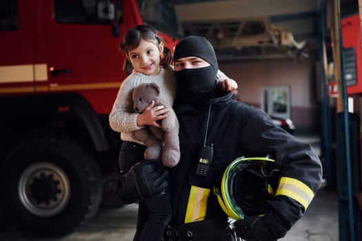 Holds teddy bear. Happy little girl is with male firefighter in protective uniform.