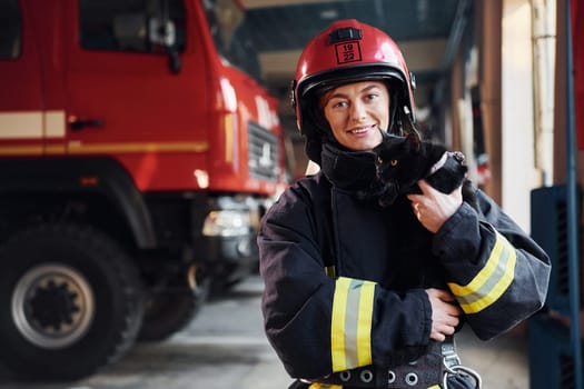 Cute black cat. Female firefighter in protective uniform standing near truck.
