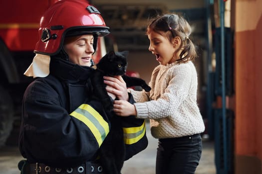 Cute black cat. Happy little girl is with female firefighter in protective uniform.