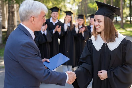 The teacher shakes hands with the student and presents the diploma outdoors. A group of university graduates