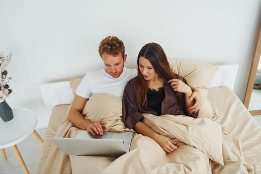 Couple lying down on bed with laptop. Interior and design of beautiful modern bedroom at daytime.