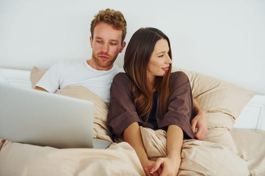 Couple lying down on bed with laptop. Interior and design of beautiful modern bedroom at daytime.