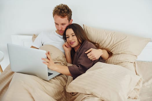 Couple lying down on bed with laptop. Interior and design of beautiful modern bedroom at daytime.