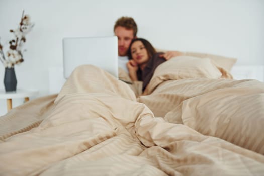 Couple lying down on bed with laptop. Interior and design of beautiful modern bedroom at daytime.