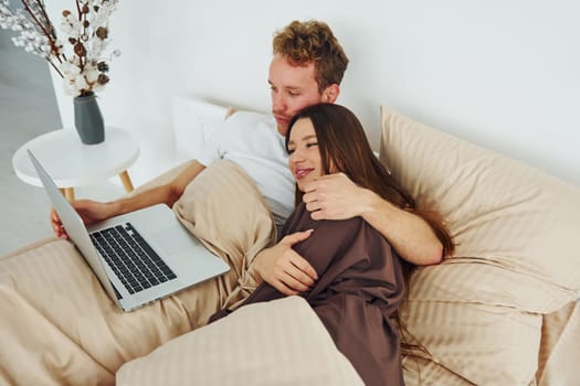 Couple lying down on bed with laptop. Interior and design of beautiful modern bedroom at daytime.