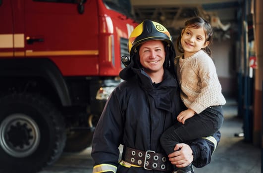 Happy little girl is with male firefighter in protective uniform.