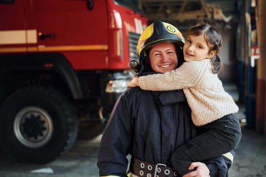 Happy little girl is with male firefighter in protective uniform.