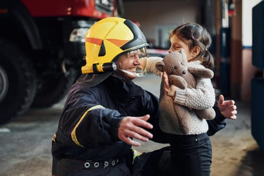 Happy little girl is with male firefighter in protective uniform.