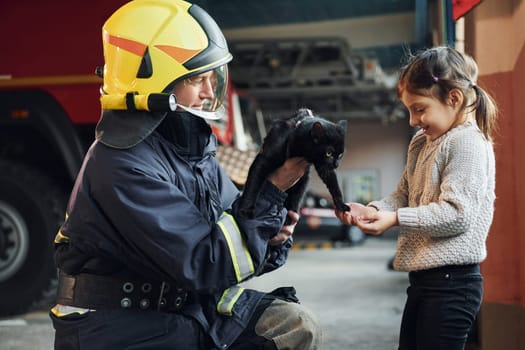 Cute black cat. Happy little girl is with male firefighter in protective uniform.