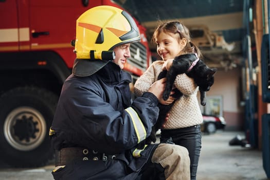 Cute black cat. Happy little girl is with male firefighter in protective uniform.
