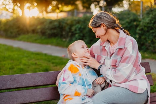 mother and son sit on a park bench in the rays of the setting sun. the concept of a family. Mother's Day. beautiful girl (mother) with a boy (son) in the park in the park are sitting on a bench at sunset.