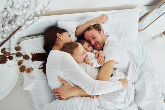 Woman and man lying down on bed with their children. Interior and design of beautiful modern bedroom at daytime.
