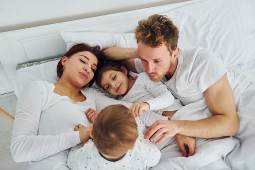 Woman and man lying down on bed with their children. Interior and design of beautiful modern bedroom at daytime.