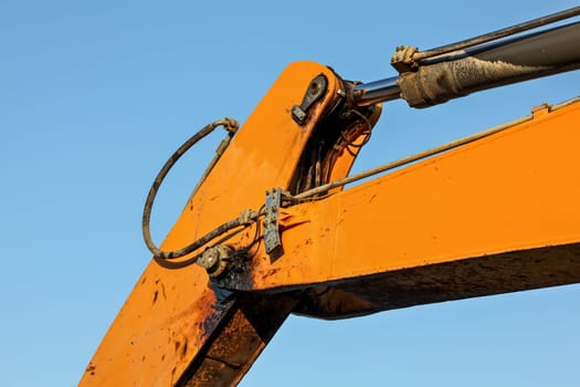 Detail on orange arm of excavator machine, hydraulic piston mechanism and joint dirty from black oil visible. Afternoon blue sky contrast in background.