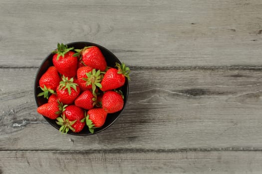 Tabletop view - small black ceramic bowl with freshly picked strawberries, on gray wood desk. Space for text on the right.