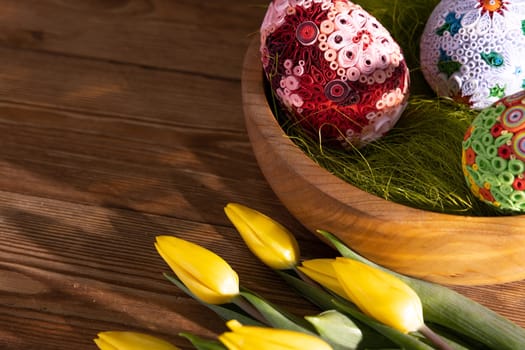 Patterned Easter eggs arranged on hay in a wooden bowl stand on a wooden table top. A view of the Easter composition as seen from above.