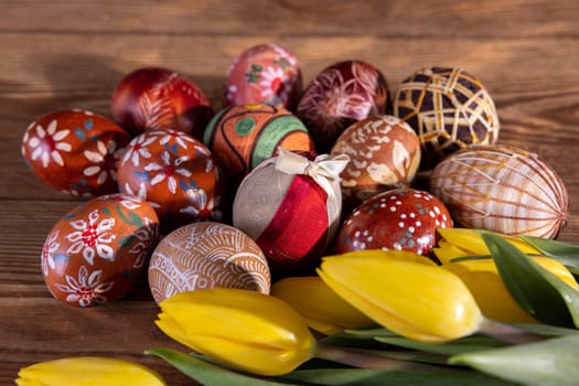 Easter eggs with different patterns kept in brown colors lie side by side on the boards and yellow tulips highlight the Easter composition.