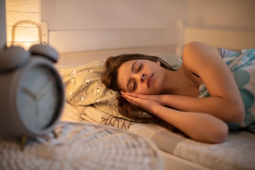 The sleeping young woman smiles slightly in her sleep. A girl under a soft blanket. A large alarm clock stands on the bedside table.