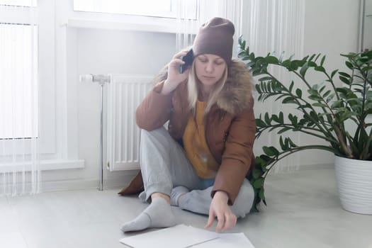 The woman is warmly dressed in a brown jacket and hat, sitting near the heater with bills in her hands and talking on the phone. The concept of a crisis and a large set for utilities