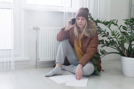 The woman is warmly dressed in a brown jacket and hat, sitting near the heater with bills in her hands and talking on the phone. The concept of a crisis and a large set for utilities