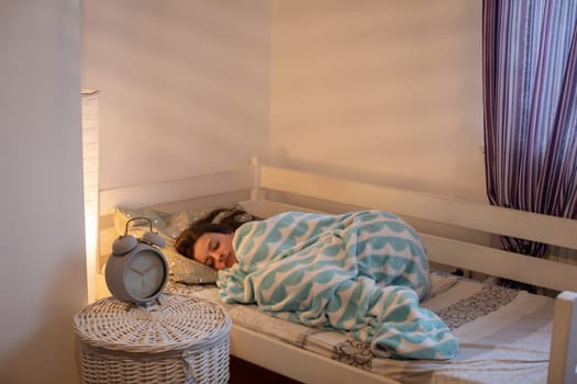 A curled-up woman lies on her bed wrapped in a blue blanket. The light of a lamp on the wall.