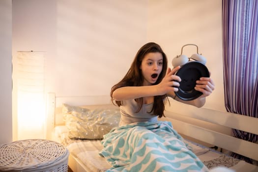 A sleep-deprived girl looks surprised at her alarm clock. The light of a lamp on the wall.